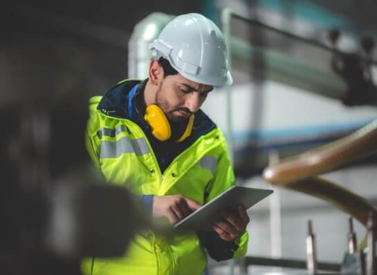 Service technician wearing a hard hat, high-visibility vest, and using a tablet while working on an industrial facility