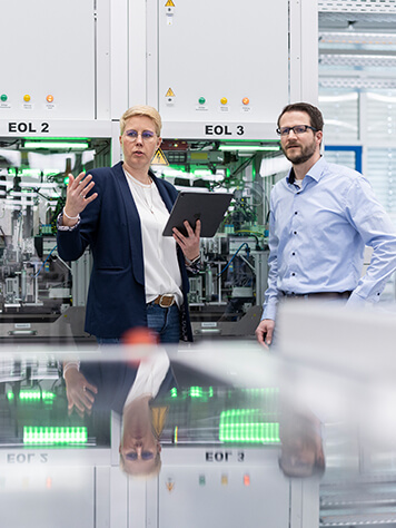 A man and a woman stand in front of a control cabinet with a tablet and look into the factory. Representation of cyber security.​