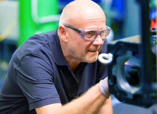A man wearing safety goggles is intently examining a mechanical component in a workshop