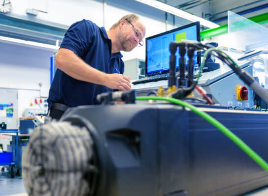 A man wearing glasses and work clothes is standing next to a machine with a monitor and several cables