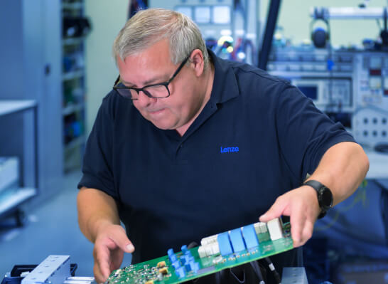 A man wearing glasses is intently examining an electronic circuit board in a workshop.