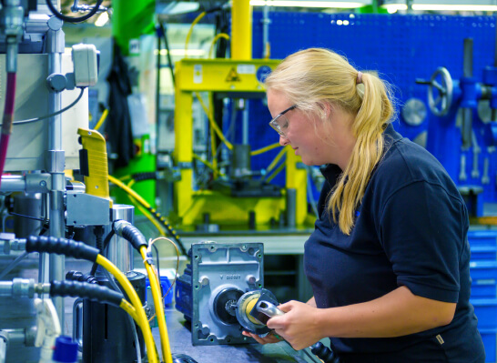 Frau h&auml;lt konzentriert ein mechanisches Bauteil in einer Produktionshalle mit Maschinen im Hintergrund.