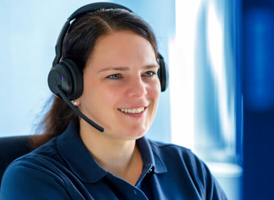 A woman wearing a headset smiles warmly at the camera while working at a computer screen in an office.
