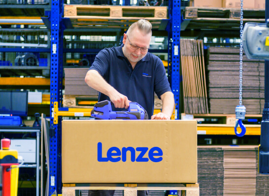 A man carefully packs a box labeled "Lenze" in a warehouse with shelves full of boxes.