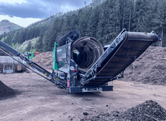 Large machine with two wide conveyor belts and a cylindrical drum screen on a construction site in front of a forest